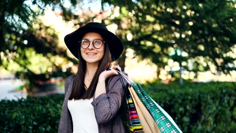 Porträt-der-gut-aussehende-Mädchen-mit-Hut-und-Brille-hält-Einkaufstaschen-Blick-in-die-Kamera-und-lächelnd.-Elegante-Menschen,-Einkauf-und-Jugend-Konzept.
