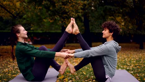 Chicas-atractivas-están-haciendo-ejercicios-de-yoga-en-pareja,-cogidos-de-la-mano-y-poner-pies-juntos-elevando-las-piernas-sentado-en-la-estera-en-el-parque.-Concepto-de-entrenamiento-y-gente-agradable.