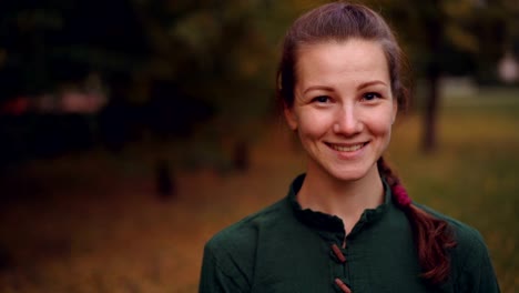 Close-up-portrait-of-beautiful-girl-with-braid-looking-at-camera-and-smiling-then-laughing-standing-outdoors-in-park-in-autumn-with-trees-in-background.