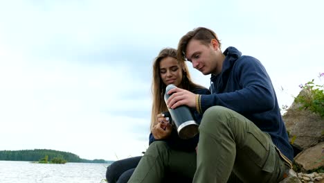 Un-chico-con-una-chica-está-sentada-en-la-orilla-del-río-y-bebiendo-té-de-un-termo.