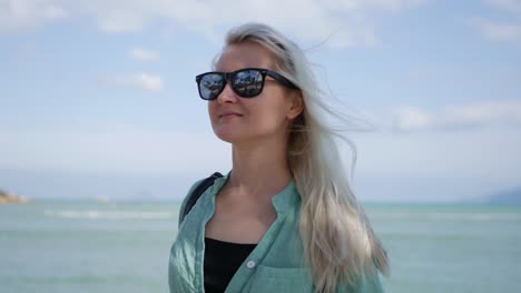 Closeup-of-young-happy-caucasian-woman-with-long-blonde-hair-in-sunglasses-and-green-shirt-standing-and-smiling-near-palm-tree-on-a-blue-sea-background.-Travel-concept