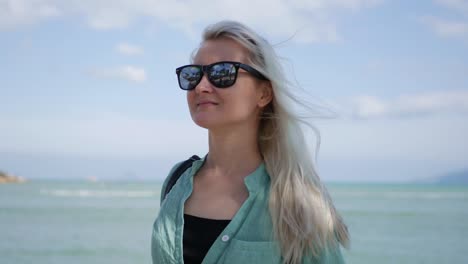 Primer-plano-de-mujer-caucásica-feliz-joven-con-cabello-largo-Rubio-en-gafas-de-sol-y-camiseta-verde-de-pie-y-sonriendo-junto-a-la-palmera-sobre-un-fondo-azul-del-mar.-Concepto-de-viaje