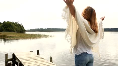 Young-woman-relaxes-on-lake-pier,-stands-arms-outstretched