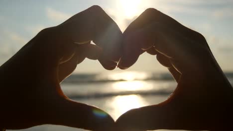 Muchacha-haciendo-el-corazón-con-sus-manos-sobre-fondo-del-mar-con-hermosa-puesta-de-sol-de-oro.-Silueta-de-mujer-brazo-en-forma-de-corazón-con-salida-del-sol-dentro.-Concepto-de-vacaciones.-Vacaciones-de-verano-en-la-playa