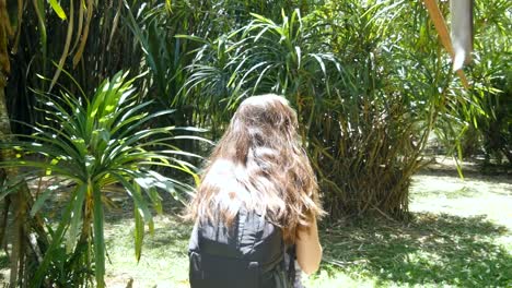 Happy-girl-with-backpack-is-walking-along-green-jungle-and-having-fun.-Young-beautiful-woman-going-and-spinning-at-tropical-forest.-Freedom-travel-concept.-Rear-back-view