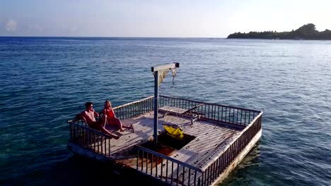 v03845-Aerial-flying-drone-view-of-Maldives-white-sandy-beach-2-people-young-couple-man-woman-relaxing-on-sunny-tropical-paradise-island-with-aqua-blue-sky-sea-water-ocean-4k-floating-pontoon-jetty-sunbathing-together