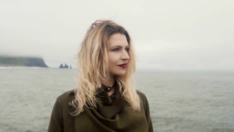 Young-blonde-woman-standing-on-black-beach,-near-the-Troll-toes-in-Iceland-and-looking-around,-hair-waving-on-the-wind