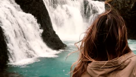 Close-up-view-of-young-attractive-woman-standing-alone-and-looking-on-powerful-waterfall-in-Iceland,-hair-wave-on-wind