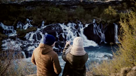 Back-view-of-young-traveling-couple-standing-in-mountains-near-the-waterfalls-in-Iceland-and-taking-photo-on-smartphone