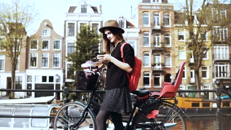 Blogger-lady-with-bicycle-on-an-old-town-bridge.-Female-European-tourist-types,-looks-around-enjoying-warm-sunny-day.-4K