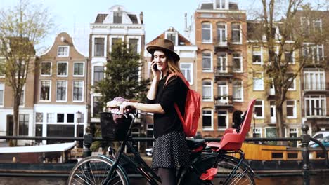 Smiling-lady-talking-on-the-phone,-cycling-away.-Young-businesswoman-on-a-magnificent-bridge.-Amsterdam,-Netherlands.-4K