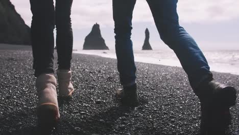 Cerrar-tiro-de-joven-pareja-cogidos-de-la-mano-corriendo-por-la-playa-de-arena-negra-en-Islandia,-cámara-lenta