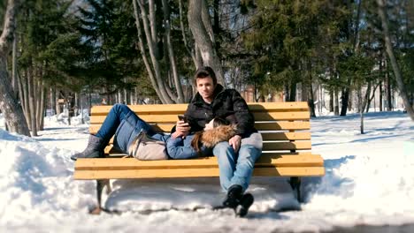 Frau-schläft,-Mann-betrachten-Telefon.-Mann-und-Frau-ausruhen-zusammen-im-Winter-City-Park-auf-einer-Bank.-Sonnigen-Wintertag.