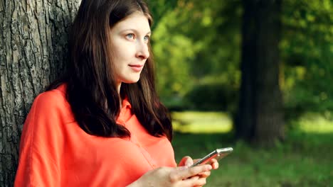 Beautiful-young-lady-is-using-smartphone-resting-in-park-under-tree-and-enjoying-modern-technology-and-summer-nature.-People,-summertime-and-communication-concept.