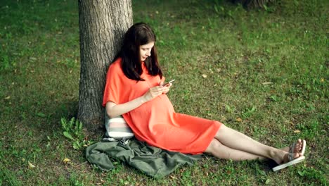 Relaxed-pregnant-girl-is-using-smartphone-touching-screen-sitting-on-grass-in-park-on-warm-summer-day.-Pregnancy,-modern-technology-and-nature-concept.