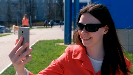 Portrait-of-happy-young-brunette-woman-in-sunglasses-talking-on-video-calling-on-the-phone-beside-blue-building-on-the-street.