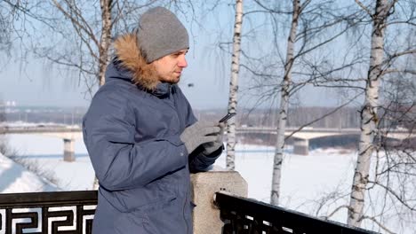 Hombre-en-azul-por-la-chaqueta-con-capucha-de-piel-utilizando-su-teléfono-celular-para-web-y-charlando-en-un-parque-de-invierno.