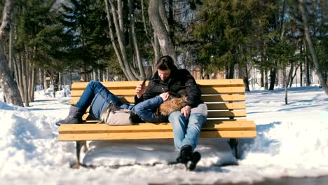 Mann-und-Frau-ausruhen-zusammen-im-Winter-City-Park-auf-einer-Bank.-Sonnigen-Wintertag.-Suchen-Sie-etwas-auf-dem-Handy.