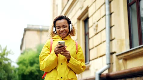 Dolly-shot-of-cheerful-young-lady-listening-to-music-with-headphones,-singing-and-using-smartphone-walking-outdoors-in-modern-city.-Fun,-people-and-technology-concept.