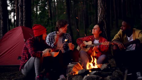 Happy-men-and-women-are-sitting-around-fire-cooking-marshmallow-playing-the-guitar-and-singing-during-hike-in-forest.-Young-people-are-having-fun-and-enjoying-nature.