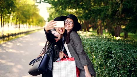 Excited-female-friends-are-taking-selfie-using-smartphone-and-holding-shopping-bags-with-purchases-on-sunny-autumn-day.-Modern-technology-and-youth-concept.