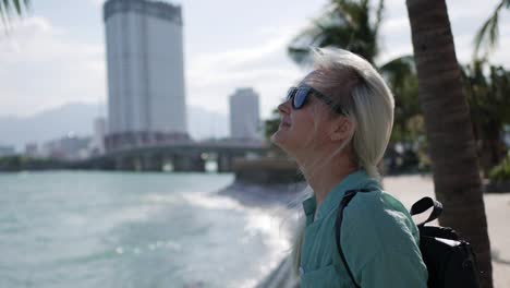 Young-happy-caucasian-woman-with-long-blonde-hair-in-sunglasses-and-green-shirt-standing-and-smiling-near-palm-tree-on-a-park-and-sea-background.-Travel-concept