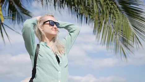 Young-happy-caucasian-woman-with-long-blonde-hair-in-sunglasses-and-green-shirt-standing-and-smiling-near-palm-tree-on-a-blue-sky-background.-Travel-concept