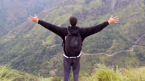 Young-man-tourist-with-backpack-standing-on-the-edge-of-beautiful-canyon-and-victoriously-raised-hands.-Male-hiker-reaching-up-top-of-mountain-and-outstretching-arms-up.-Rear-back-view