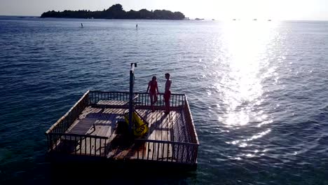 v03831-Aerial-flying-drone-view-of-Maldives-white-sandy-beach-2-people-young-couple-man-woman-relaxing-on-sunny-tropical-paradise-island-with-aqua-blue-sky-sea-water-ocean-4k-floating-pontoon-jetty-sunbathing-together