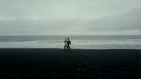 Aerial-view-of-young-couple-walking-on-black-volcanic-beach.-Man-and-woman-have-a-date-on-shore-of-the-sea-in-Iceland