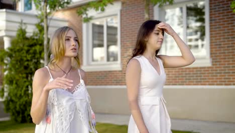 Two-young-friends-walk-next-to-brick-country-houses,-women-talk-about-life-style-and-fashion-on-a-summer-day