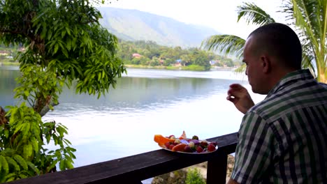 Un-hombre-está-comiendo-fruta-y-mirando-el-lago-y-las-montañas