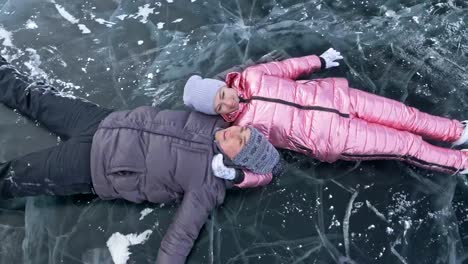 Young-couple-has-fun-during-winter-walk-against-background-of-ice-of-frozen-lake.-Lovers-lie-on-clear-ice-with-cracks,-have-fun,-kiss-and-hug.-View-from-above.-Honeymoon.-Love-story.