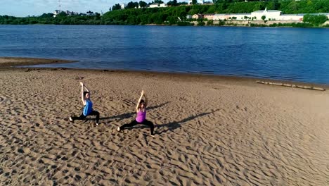 Zwei-Frau-Yoga-am-Sandstrand-am-Fluss-in-der-Stadt-zu-tun.-Blick-auf-die-schöne-Stadt-im-Sonnenaufgang.