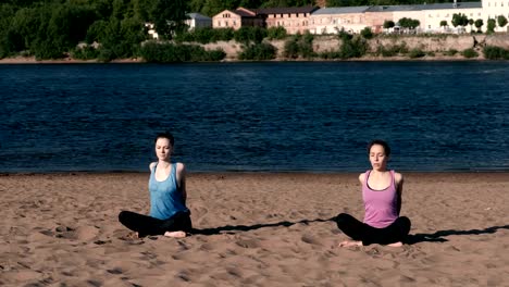 Two-woman-stretching-yoga-on-the-beach-by-the-river-in-the-city.-Beautiful-city-view.-Meditation-pose.