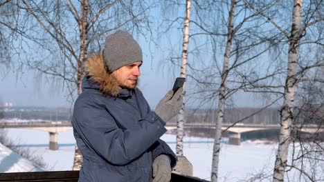 Mann-in-blau-Daunenjacke-mit-Fell-Kapuze-mit-Handy-für-Video-Chat-und-lächelnd-in-Winter-Park.