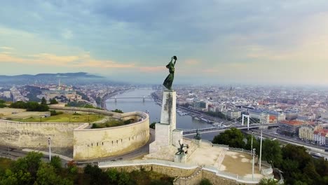 Herumfliegen-der-Statue-of-Liberty-mit-der-Skyline-von-Budapest-im-Hintergrund.-Budapest,-Ungarn---4K-Luftaufnahmen-bei-Sonnenaufgang