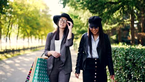 Tilt-down-toma-de-adictos-a-las-compras-feliz-Milenio-mujer-hablar-y-caminar-junto-con-bolsas-brillantes.-Las-mujeres-están-usando-gafas-y-ropa-de-moda.