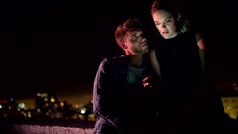 Young-couple-looking-at-smart-phone-on-rooftop-at-night