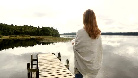 Young-adult-relaxes-on-jetty-above-lake,-uses-digital-tablet