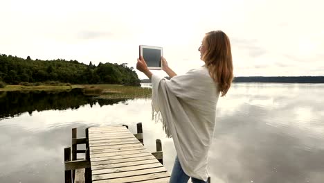 Young-adult-relaxes-on-jetty-above-lake,-uses-digital-tablet