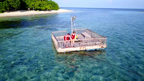 v03852-Aerial-flying-drone-view-of-Maldives-white-sandy-beach-2-people-young-couple-man-woman-relaxing-on-sunny-tropical-paradise-island-with-aqua-blue-sky-sea-water-ocean-4k-floating-pontoon-jetty-sunbathing-together