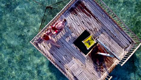 v03823-Aerial-flying-drone-view-of-Maldives-white-sandy-beach-2-people-young-couple-man-woman-relaxing-on-sunny-tropical-paradise-island-with-aqua-blue-sky-sea-water-ocean-4k-floating-pontoon-jetty-sunbathing-together