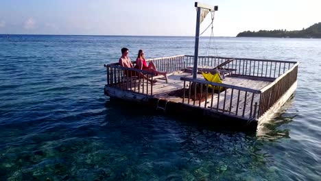 v03847-Aerial-flying-drone-view-of-Maldives-white-sandy-beach-2-people-young-couple-man-woman-relaxing-on-sunny-tropical-paradise-island-with-aqua-blue-sky-sea-water-ocean-4k-floating-pontoon-jetty-sunbathing-together