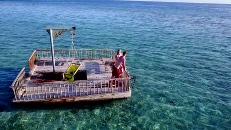 v03856-Aerial-flying-drone-view-of-Maldives-white-sandy-beach-2-people-young-couple-man-woman-relaxing-on-sunny-tropical-paradise-island-with-aqua-blue-sky-sea-water-ocean-4k-floating-pontoon-jetty-sunbathing-together