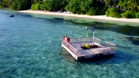 v03853-Aerial-flying-drone-view-of-Maldives-white-sandy-beach-2-people-young-couple-man-woman-relaxing-on-sunny-tropical-paradise-island-with-aqua-blue-sky-sea-water-ocean-4k-floating-pontoon-jetty-sunbathing-together