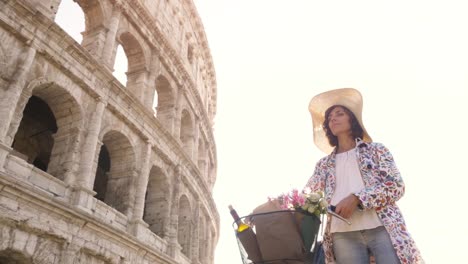 Beautiful-young-woman-in-colorful-fashion-dress-walking-alone-with-bike-arriving-in-front-of-colosseum-in-Rome-at-sunset-with-happy-attractive-tourist-girl-with-straw-hat-ground-shot