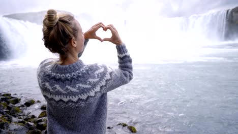 Young-woman-in-Iceland-making-heart-shape-finger-frame-on-spectacular-waterfall