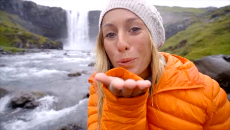 Junge-Frau-unter-Selfie-Porträt-mit-herrlichen-Wasserfall-in-Island,-weht-einen-Kuss-auf-Kamera.-Die-Leute-reisen-Exploration-Konzept