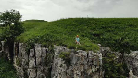 Young-hipster-woman-is-sitting-on-the-edge-on-the-grass-of-tall-wall-of-rock.-Aerial-view.-Drone-is-flying-slow-backward-from-model.-Establishing-revealing-shot